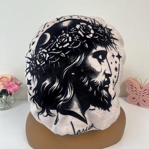 K&ouml;nnte beinhalten: Eine hellrosa Haube mit einer schwarz-wei&szlig;en Illustration einer religi&ouml;sen Figur mit einer Dornenkrone, einem Halbmond und Sternen. Das Wort "Jesus" ist in Kursivschrift auf der Haube geschrieben.