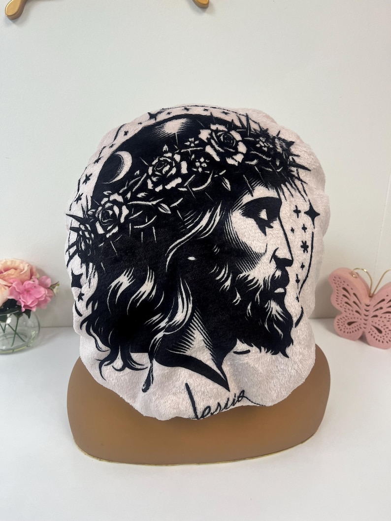 K&ouml;nnte beinhalten: Eine hellrosa Haube mit einer schwarz-wei&szlig;en Illustration einer religi&ouml;sen Figur mit einer Dornenkrone, einem Halbmond und Sternen. Das Wort "Jesus" ist in Kursivschrift auf der Haube geschrieben.