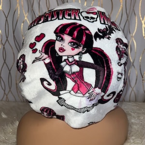 May include: White plush sleep cap featuring a Monster High character design. The cap has a repeating pattern of the character, hearts, and bats. The Monster High logo is printed across the top.