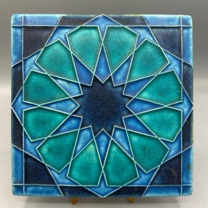 May include: Square ceramic tile with a geometric starburst design in shades of teal, turquoise, and navy blue. The tile has a glossy finish and a raised pattern, creating a textured appearance. The tile is approximately 6 inches square.