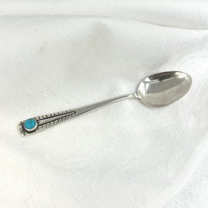 May include: A silver spoon with a turquoise stone inlay on the handle. The spoon has a rounded bowl and a decorative handle with etched details. The spoon is set against a white background.