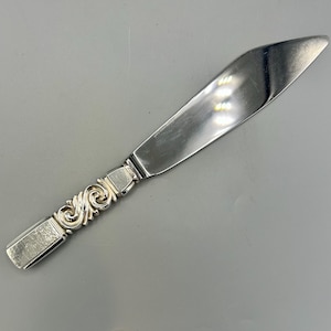 May include: A silver butter knife with a decorative handle featuring a scroll design. The blade is smooth and reflective, and the handle has a rectangular end. The knife is designed for spreading butter or other soft spreads.