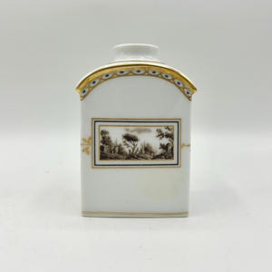 May include: A white porcelain tea caddy with a rectangular body and a curved top. The piece features a central rectangular panel with a sepia-toned landscape illustration, framed by gold and blue lines. Gold trim accents the edges and top border.
