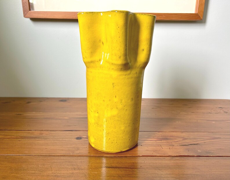 Puede incluir: Un jarr&oacute;n alto de cer&aacute;mica amarillo brillante con una parte superior ondulada &uacute;nica. El jarr&oacute;n tiene una superficie texturizada y un cuerpo cil&iacute;ndrico. Se encuentra sobre una superficie de madera, con una obra de arte enmarcada visible en el fondo.