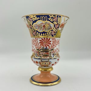 May include: A porcelain vase featuring a flared rim, a white body with intricate floral patterns in red, orange, and gold, and a deep blue band near the top. The base is a solid peach color with a gold-trimmed blue edge, standing on a pedestal.
