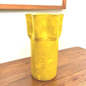 Puede incluir: Un jarr&oacute;n alto de cer&aacute;mica amarilla con borde acanalado. El jarr&oacute;n tiene un cuerpo cil&iacute;ndrico y una superficie texturizada. Est&aacute; sobre una mesa de madera. Una obra de arte enmarcada cuelga en el fondo.