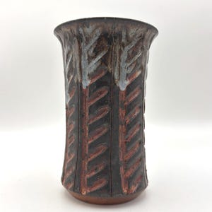 May include: A cylindrical ceramic vase with a flared rim, featuring a dark brown and black glaze with vertical, carved, leaf-like patterns in a reddish-brown hue. The upper portion of the vase shows subtle white and grey highlights.