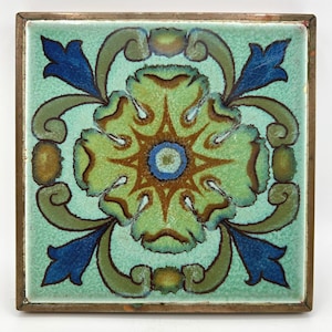 May include: A square ceramic tile featuring a central floral motif in shades of light green, olive, and brown, surrounded by dark blue leaf-like accents on a pale mint background. The tile is encased in a thin, weathered metallic frame.