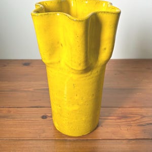 Puede incluir: Un jarr&oacute;n de cer&aacute;mica amarillo brillante con borde ondulado. El jarr&oacute;n tiene forma cil&iacute;ndrica y superficie texturizada. Se encuentra sobre una superficie de madera.
