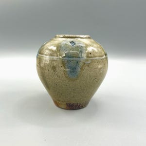 May include: A small, round ceramic vase with a speckled, earthy color palette. The vase features a light brown base, transitioning to a tan body with blue accents near the top. The rim is a lighter shade, and the vase has a textured surface.