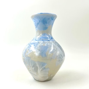 May include: A ceramic vase with a bulbous body and a flared rim, featuring a smooth, light beige base glaze decorated with crystalline patterns in shades of blue and white that radiate across the surface of the vessel in floral-like shapes.
