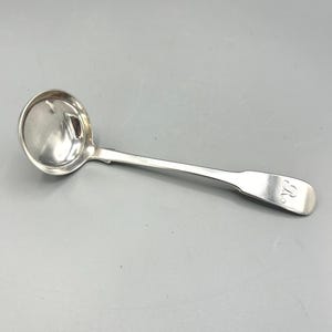 May include: A polished silver-toned metal ladle featuring a deep, rounded bowl and a long, slender handle. The end of the handle is slightly widened and features an engraved cursive letter R, providing a distinct detail on the smooth surface.