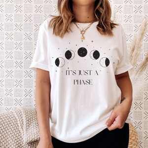 Puede incluir: Camiseta blanca con un gráfico negro de una luna creciente y estrellas. El texto "It's just a phase" está impreso debajo del gráfico.