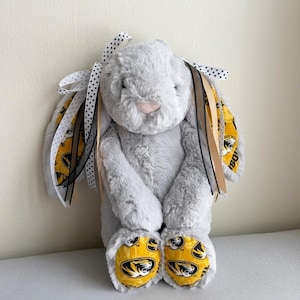 May include: A plush gray bunny with a pink nose and paws featuring a yellow and black design. The bunny's ears are adorned with ribbons in black, white, and gold. A cute stuffed animal.