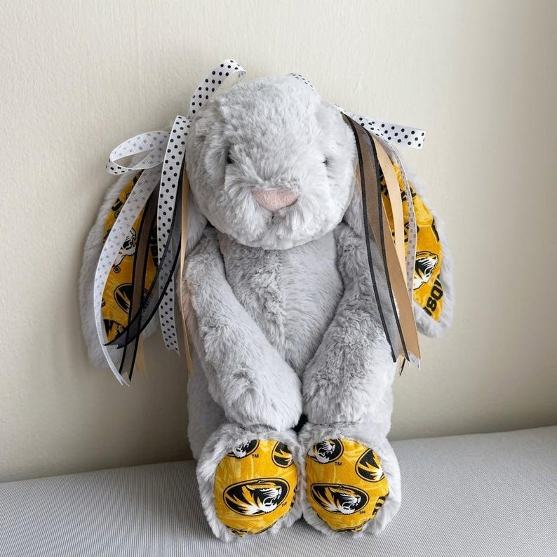 May include: A plush gray bunny with a pink nose and paws featuring a yellow and black design. The bunny's ears are adorned with ribbons in black, white, and gold. A cute stuffed animal.