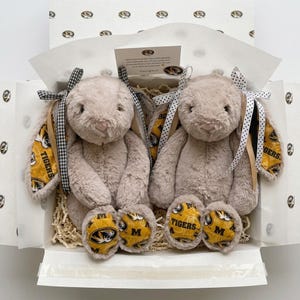 May include: Two plush bunny rabbits in a gift box. The bunnies are light brown with long ears, black and white checkered bows, and yellow and black patterned ear lining and foot pads. The box is white with a patterned interior.