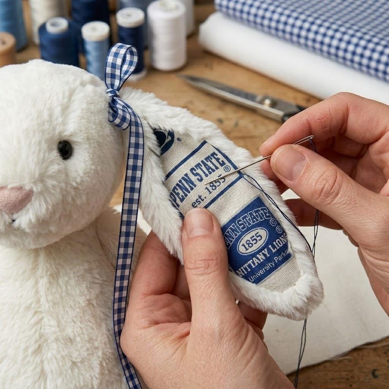 May include: A white plush bunny with a blue and white gingham ribbon tied to its ear. The bunny's ear has a sewn-on tag with the text "PENN STATE" and "1855". A needle and thread are used to sew the tag.