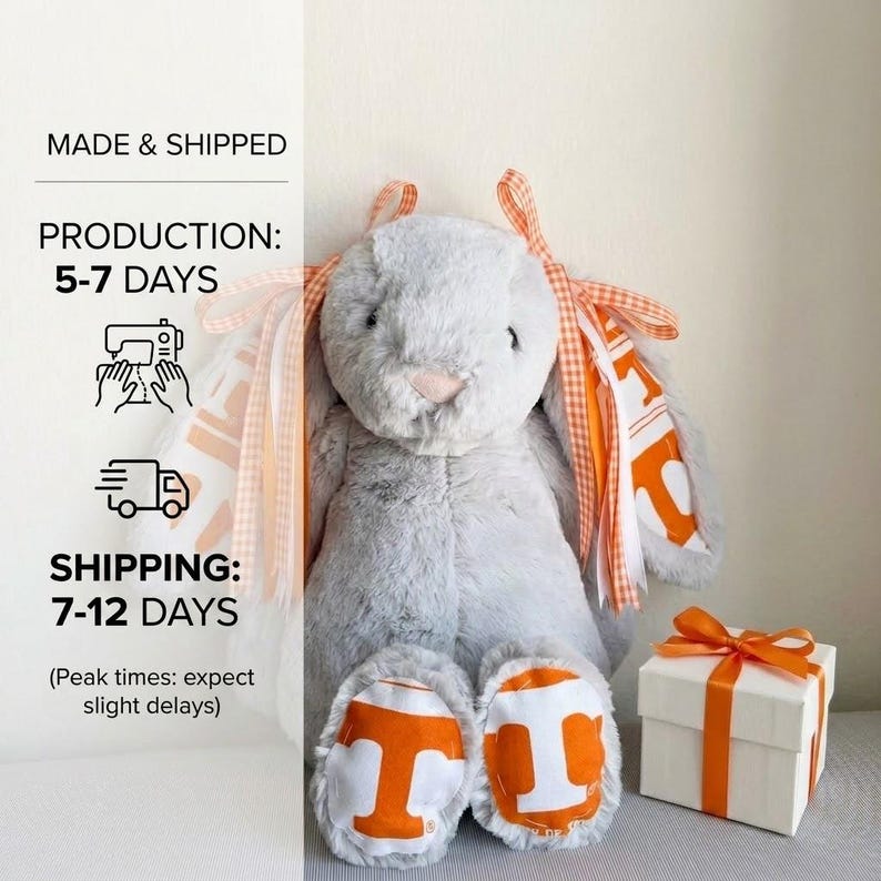 May include: A plush gray bunny with orange and white accents, including ribbons and foot pads with a large "T" logo. A white gift box with an orange ribbon sits nearby. The text "MADE & SHIPPED" and "PRODUCTION: 5-7 DAYS" is visible.