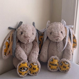 May include: Two plush bunny rabbits with tan fur, sitting side-by-side. The ears are lined with yellow fabric featuring black logos and text. Each bunny has a bow on its head and on its ears. The feet are yellow with black logos and text.