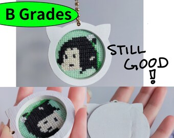 May include: White wooden keychain with a cat-shaped frame. The frame contains a green and black cross-stitch design of a person with black hair. The text "STILL GOOD!" is written in black marker on the white background.