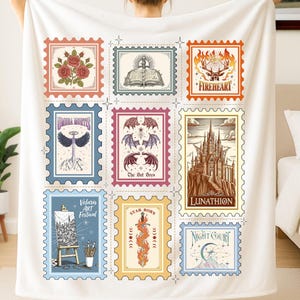 May include: A white blanket with nine colourful postage stamp designs. Each stamp features illustrations such as roses, an open book, a fiery heart, and a castle. The blanket is held up by a person.