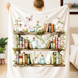 May include: White throw blanket with a watercolour illustration of a bookshelf. The design features books, bottles, flowers, and butterflies. The three wooden shelves are filled with colourful books and decorative elements.