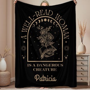 May include: Black blanket with a gold graphic that reads "A WELL-READ WOMAN IS A DANGEROUS CREATURE" with a floral and book design. The name "Patricia" is printed below the text. The blanket is held up by a person.