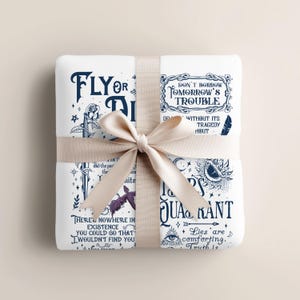 Op de afbeelding: Een witte deken met donkerblauwe tekst en illustraties, vastgebonden met een cr&egrave;mekleurig lint. De deken toont citaten en illustraties, waaronder een roos, een oog en een vleermuis. De tekst bevat zinnen als "Fly or Die" en "Don't Borrow Tomorrow's Trouble."