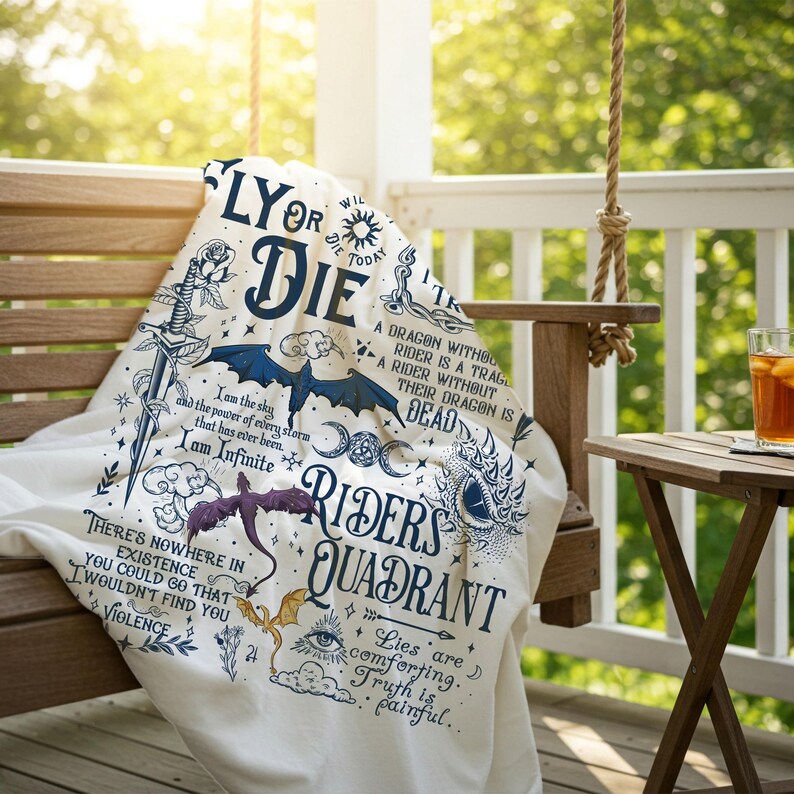 Op de afbeelding: Een witte deken met fantasie-illustraties en tekst. De deken toont draken, zwaarden en zinnen als "Fly or Die" en "Riders Quadrant" in blauwe en zwarte inkt. Het is gedrapeerd over een houten schommel.