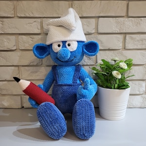 May include: Blue crocheted plush toy with a white crocheted hat, holding a red pencil. The toy is sitting on a white surface.