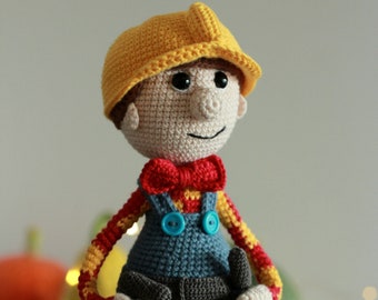 Puede incluir: Una figura de juguete de crochet que lleva un casco amarillo, una camisa a rayas rojas y amarillas, un mono azul y una pajarita roja. El juguete lleva una herramienta en cada mano.