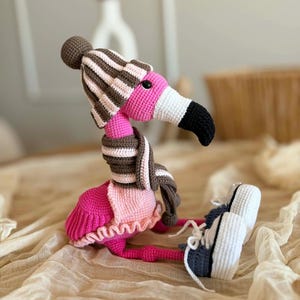 May include: A handmade crochet flamingo doll wearing a striped brown and white hat, scarf, and sneakers. The flamingo has a pink body, a white and black beak, and is sitting down. The doll is a whimsical and unique decorative item.