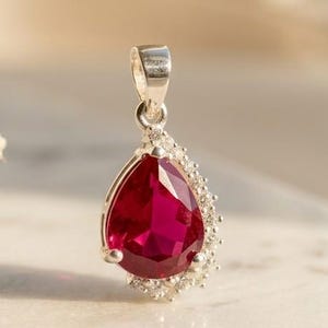 May include: A silver pendant featuring a large, pear-shaped, deep red gemstone as the centerpiece. The stone is accented by a cluster of small, clear, sparkling stones along one side, set in a polished silver frame with a sturdy bail at the top.