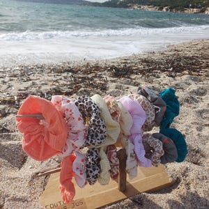 Könnte beinhalten: Ein Holzständer an einem Sandstrand hält eine Sammlung bunter Haargummis. Die Haargummis weisen verschiedene Muster und Farben auf, darunter Koralle, Blumenmuster, Leopardenmuster und Türkis. Der Text "COUTURE PUR JUS" ist auf dem Ständer zu sehen.