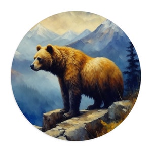 May include: Round art print featuring a majestic brown bear standing on a rocky ledge, overlooking a mountain range. The bear has thick fur and is set against a backdrop of blue and yellow hues, creating a serene landscape scene.