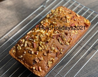 May include: A loaf of pumpkin bread topped with pecans on a wire rack. The bread is golden brown and has a slightly crispy crust. The pecans are scattered evenly over the top of the bread. The text "Hello Holidays 2021" is written on the bread.