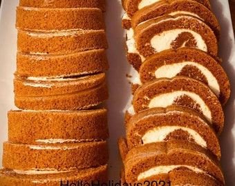May include: A platter of sliced pumpkin roll cake with a creamy white filling. The cake slices are a warm brown color, and the filling is visible in the center of each slice. The cake is arranged on a white rectangular platter.