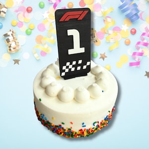 May include: A round white cake decorated with colorful sprinkles and white frosting. A black sign with the number 1 and a checkered flag design is on top of the cake. The cake is surrounded by colorful confetti and streamers.