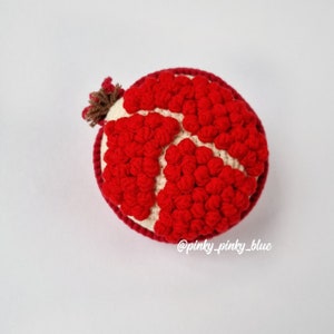 May include: A crocheted pomegranate with a red and white color scheme. The pomegranate is made of individual red balls that are stitched together to form the shape of a pomegranate. The top of the pomegranate has a brown stem.