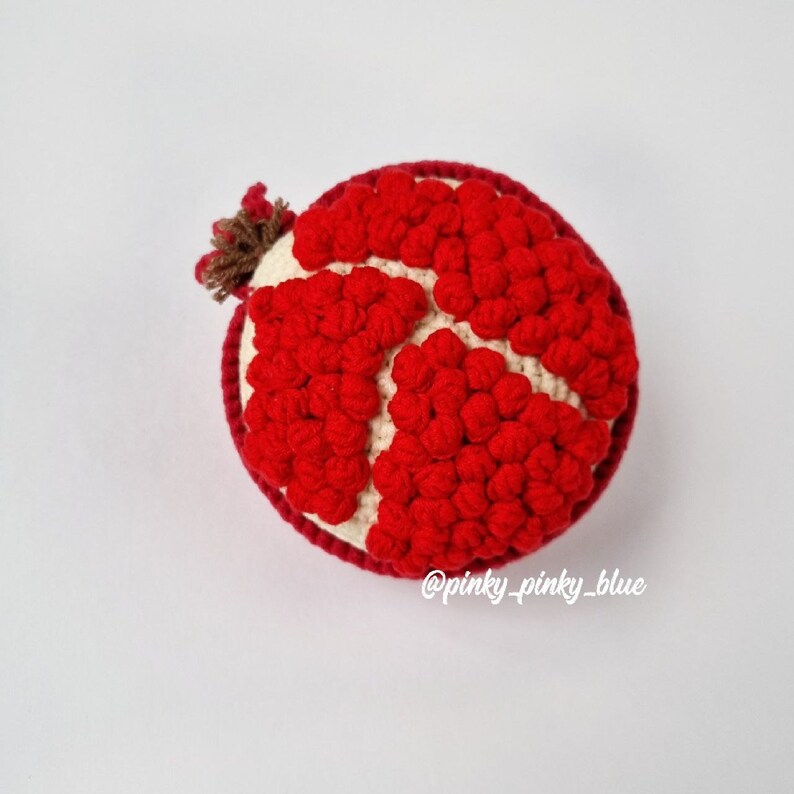 May include: A crocheted pomegranate with a red and white color scheme. The pomegranate is made of individual red balls that are stitched together to form the shape of a pomegranate. The top of the pomegranate has a brown stem.