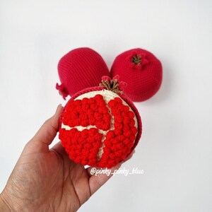 May include: Three crocheted pomegranates, one with a red interior showing, the other two with a red exterior. The pomegranate with the red interior is being held in a hand.