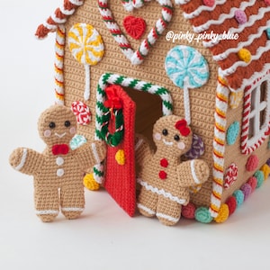 May include: A crocheted gingerbread house with candy cane trim and a red door. Two crocheted gingerbread people stand outside the house. The house is decorated with colorful candies and a wreath on the door. The text '@pinky_pinky_blue' is visible on the house.