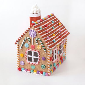 May include: A crocheted gingerbread house with a white frosting roof, candy cane trim, and colorful candy decorations. The house has a red chimney with a white puff of smoke.