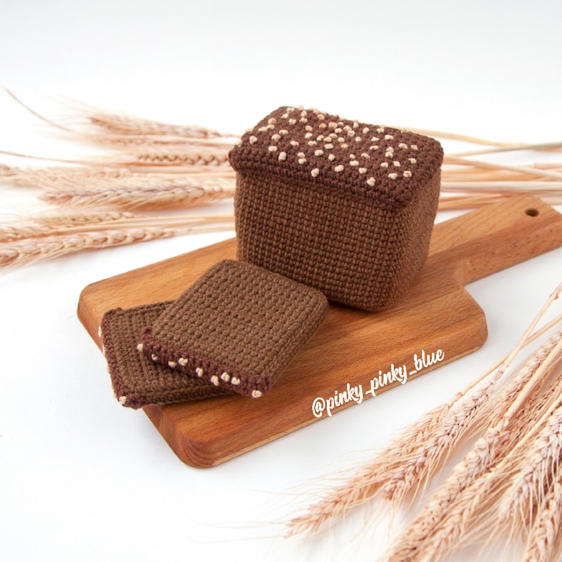 Op de afbeelding: Een gehaakt bruin brood met witte besprenkeling erop, en twee kleinere gehaakte broodsneden op een houten snijplank. De plank is omringd door tarwehalmen.