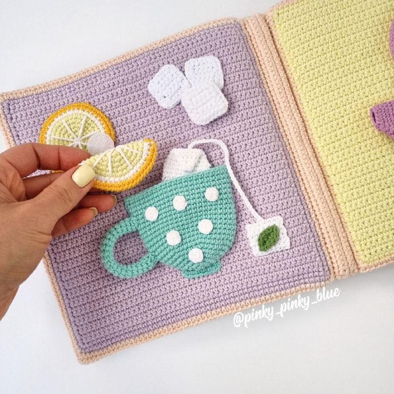 May include: A crocheted book with a teacup, lemon slices, tea bag, and a white crocheted square. The book is open and the pages are a light purple and yellow color. The text "@pinky_pinky_blue" is on the bottom right corner of the book.