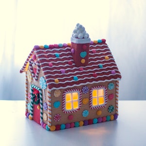 May include: A crocheted gingerbread house with candy details, a white crocheted chimney, and lit windows. The house is decorated with colorful candies and has a red door with a green wreath.