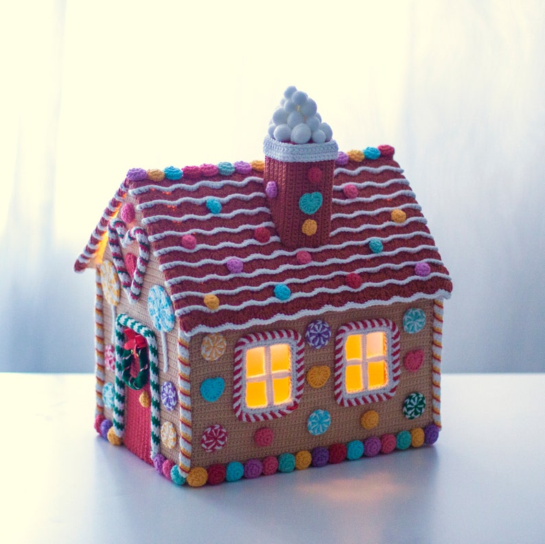 May include: A crocheted gingerbread house with candy details, a white crocheted chimney, and lit windows. The house is decorated with colorful candies and has a red door with a green wreath.
