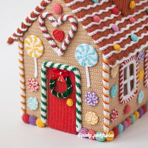 May include: A crocheted gingerbread house with a red door, candy cane trim, and a green wreath. The roof is brown with white and colorful sprinkles. The house is decorated with colorful candy and lollipops. The text "@pinky_pinky_blue" is at the bottom of the image.