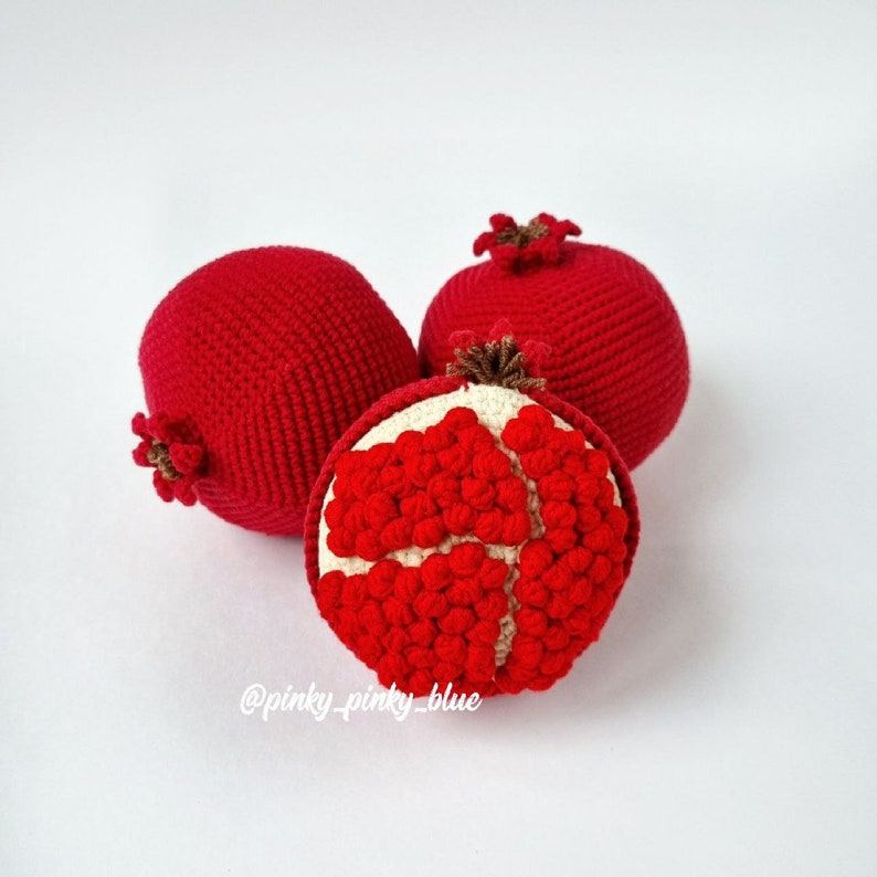 May include: Three crocheted pomegranates, one whole and two cut open to reveal the red seeds inside. The pomegranates are a bright red color and have a textured surface.
