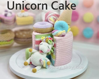 Op de afbeelding: Een gehaakte eenhoorncake met regenboogstrepen, roze glazuur en een eenhoorn die er bovenop ligt. De cake is versierd met een donut, een kegel en een snoepstok. De tekst op de cake luidt "@pinky_pinky_blue".